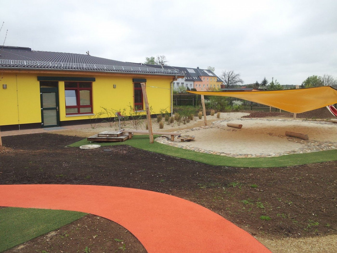 Außenanlage einer Kindereinrichtung mit gelbem Gebäude, orangefarbenem Rundkurs aus Fallschutzbelag und einem Sandbereich unter einem Sonnensegel