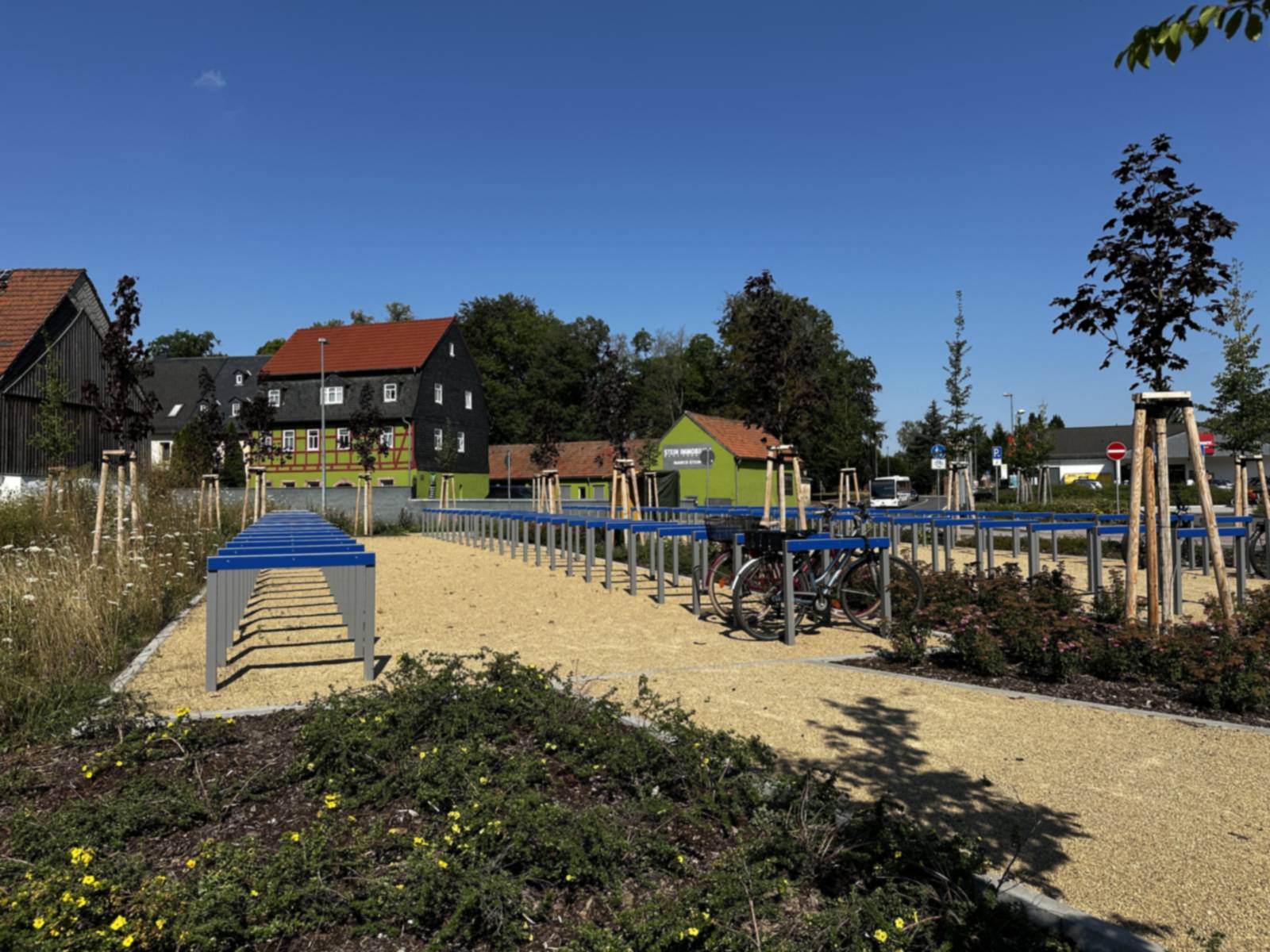 Reihen blauer Fahrradbügel auf wassergebundener Decke, davor eine Staudenpflanzung und frisch verankerte Jungbäume