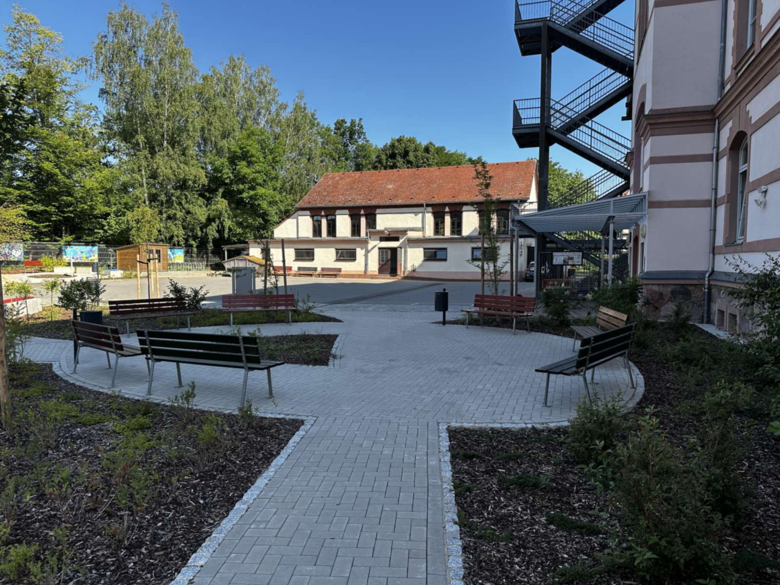 Gepflasterter Aufenthaltsbereich mit vier Bänken und Pflanzbeeten zwischen Schulgebäude und Nebengebäude