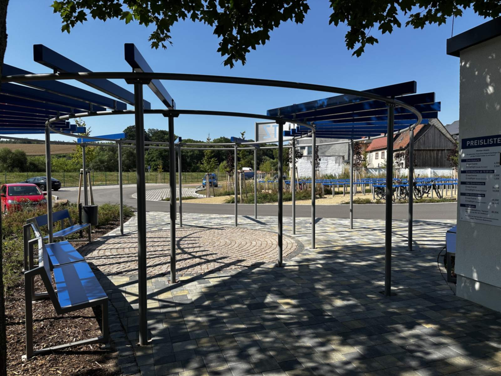 Stahlpergola über dem gepflasterten Vorplatz mit kreisförmigem Pflastermuster, links eine blaue Sitzbank