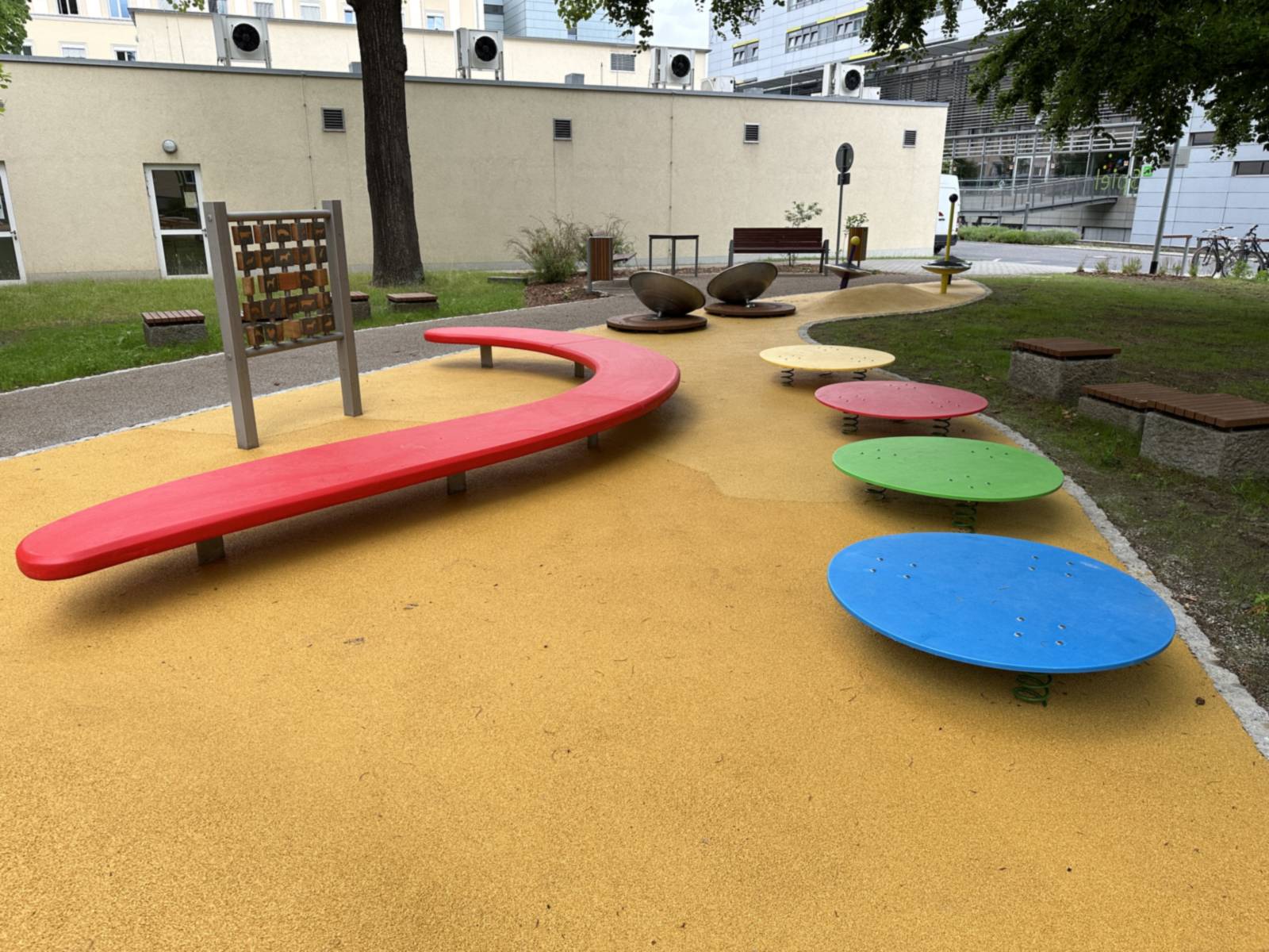 Rote Wellenbank und vier farbige Drehteller auf gelbem Fallschutzbelag, links eine Spielwand aus Holzklötzchen