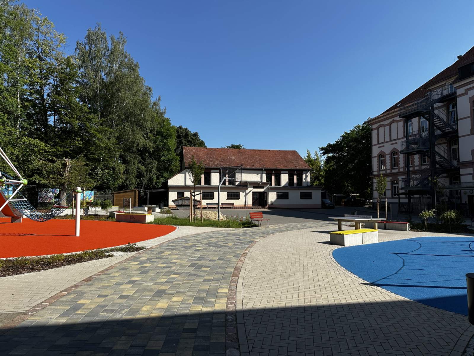 Schulhof mit rotem Fallschutzbelag und Kletteranlage links, blauem Spielbereich rechts und gepflastertem Weg dazwischen