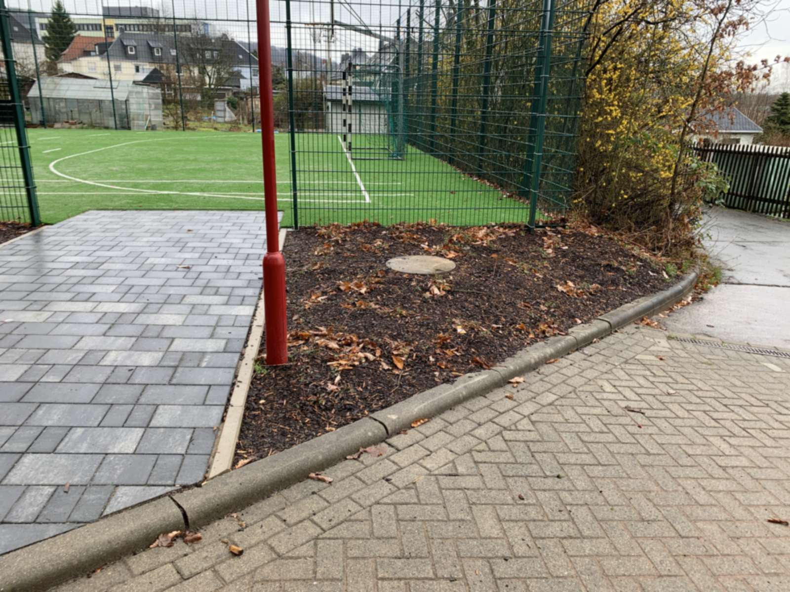 Gepflasterte Fläche mit rotem Torpfosten und Pflanzstreifen am Rand des eingezäunten Kunstrasenfelds