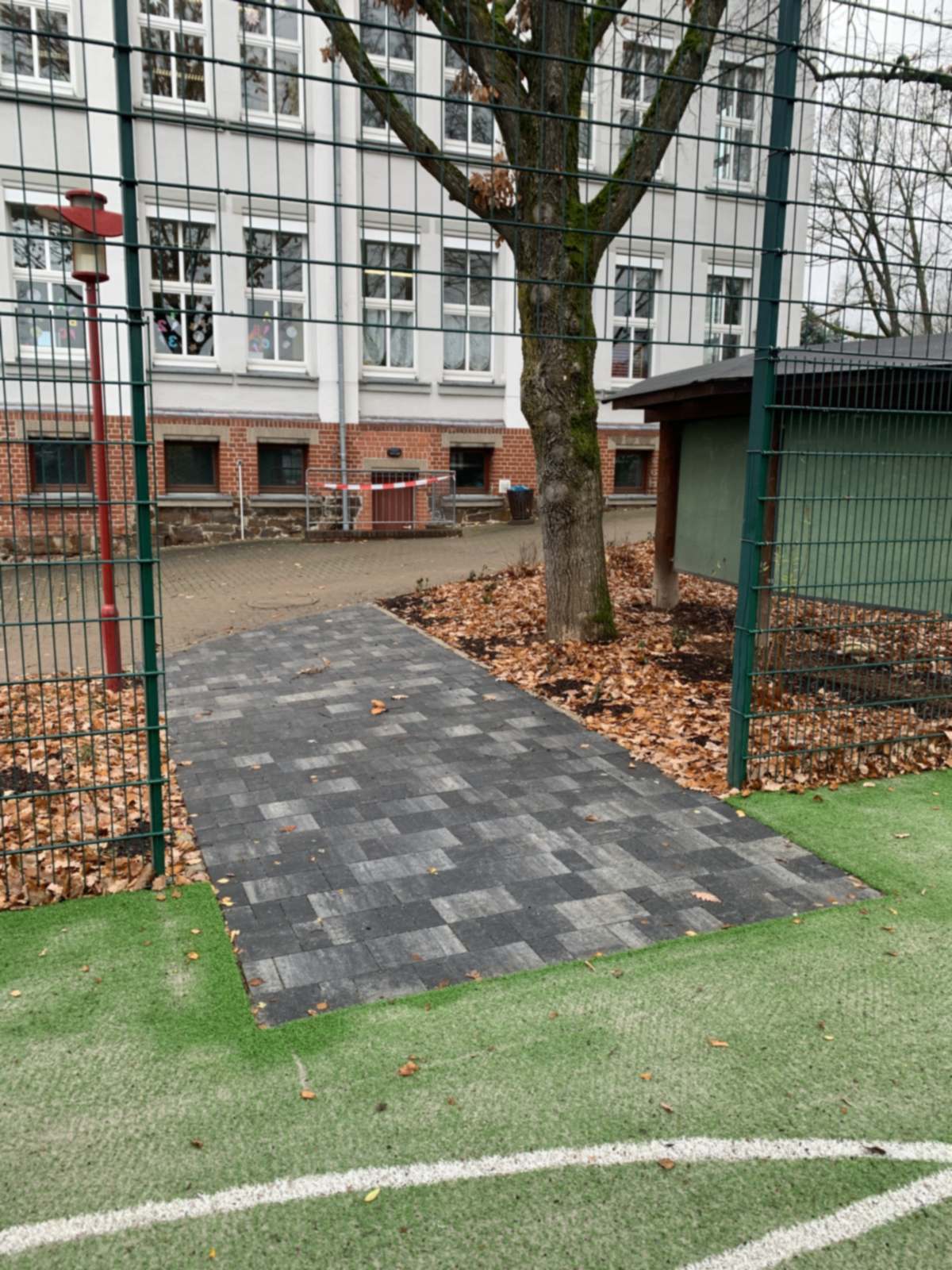 Gepflasterter Zugang vom Schulhof auf den Kunstrasen, links das Schulgebäude, rechts ein Geräteschuppen