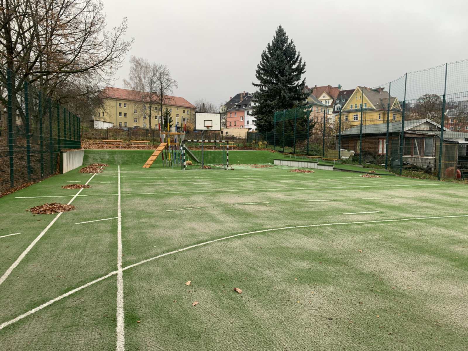 Blick über das Kunstrasen-Sportfeld mit weißer Linierung, Ballfangzaun, zwei Toren und einem Basketballkorb