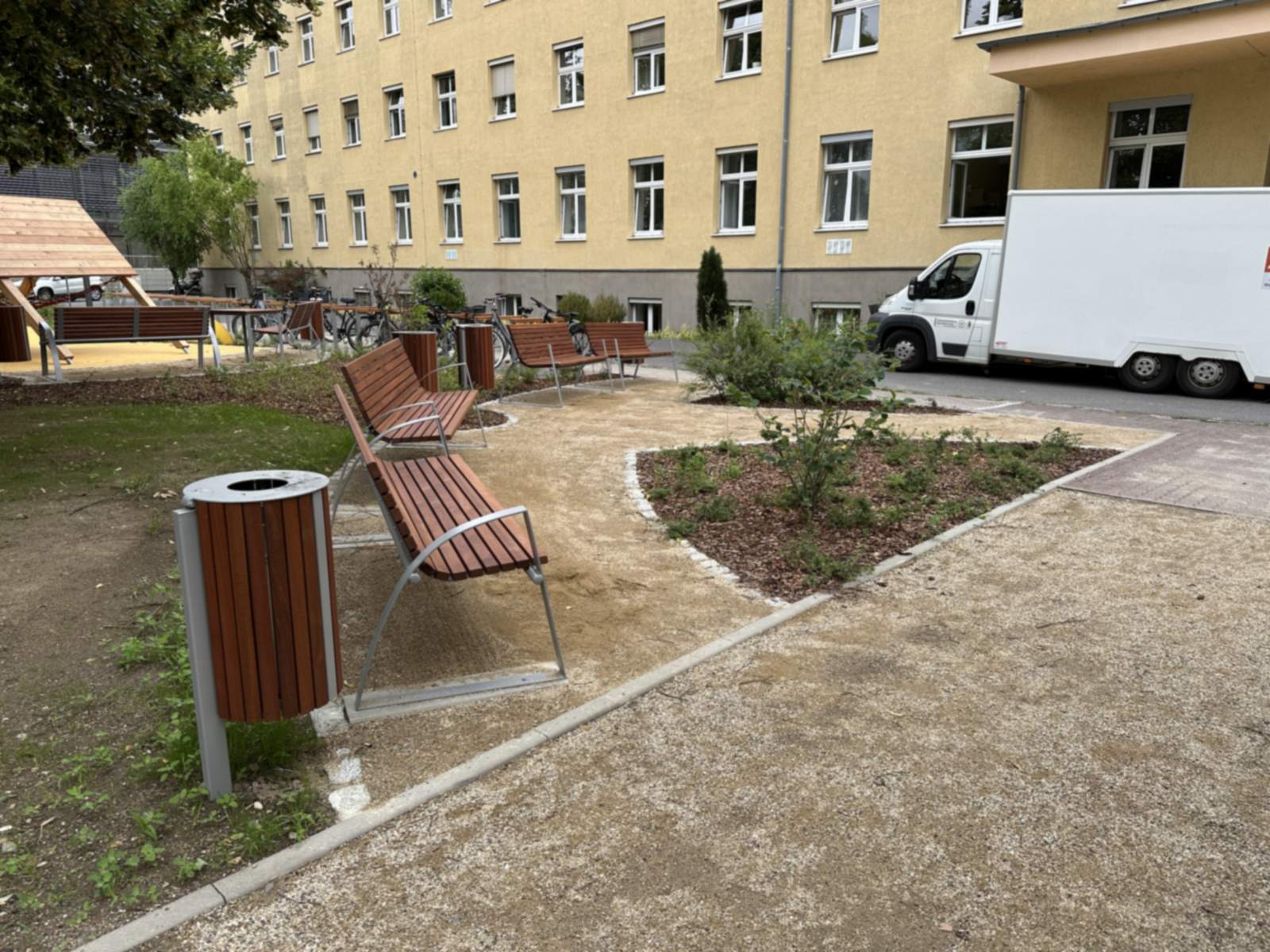 Sitzbereich mit zwei Holzbänken und Abfallbehälter an einer wassergebundenen Fläche, daneben ein neu angelegtes Staudenbeet