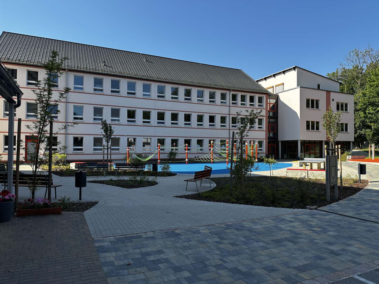 Schulhof mit blauem Spielbereich, Tischtennisplatte, Pflasterweg und jungen Bäumen
