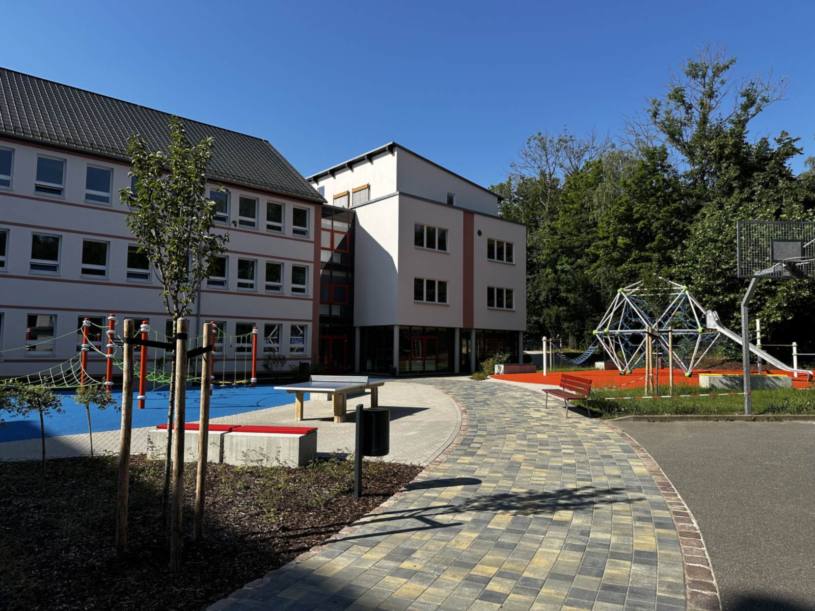 Blick über den Schulhof mit blauem Kletterbereich links und Kletterkuppel auf rotem Belag rechts