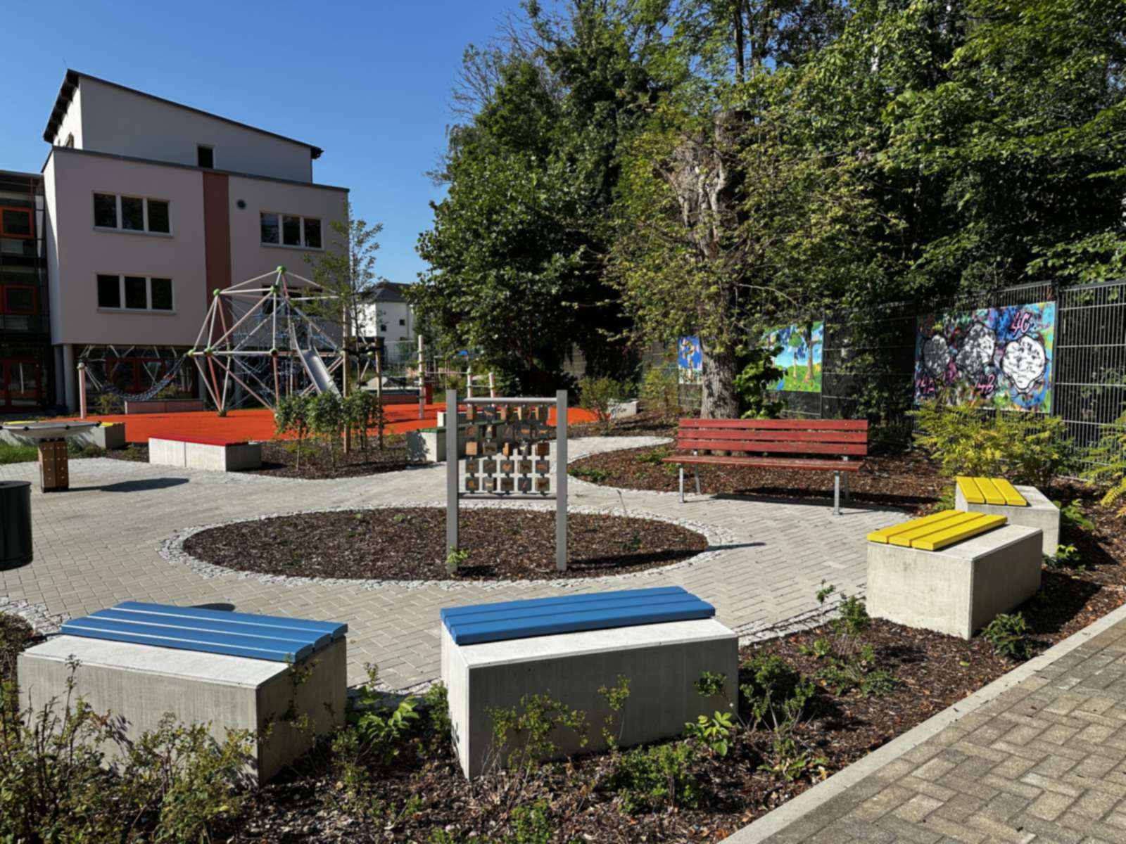 Sitzplatz mit blauen und gelben Sitzblöcken, Spielwand und Bank am Rundbeet