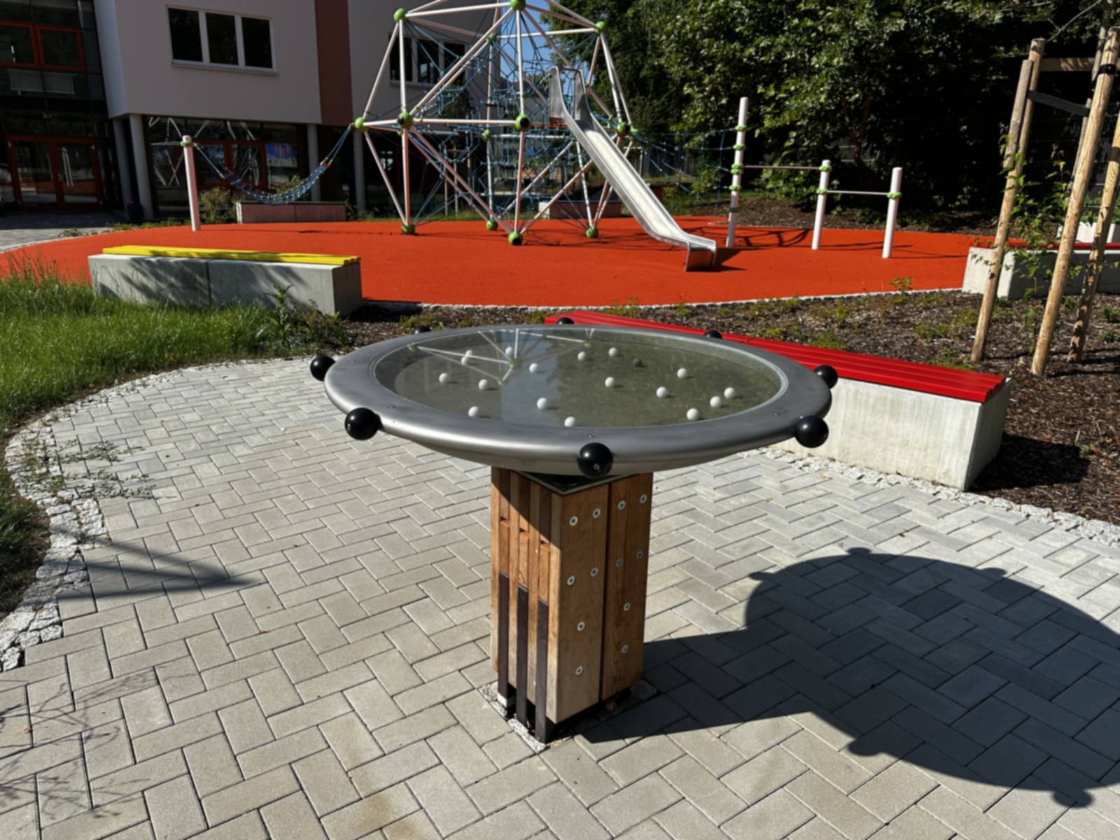 Spieltisch mit Kugelbahn auf Pflasterfläche, dahinter Kletterkuppel mit Rutsche auf rotem Belag