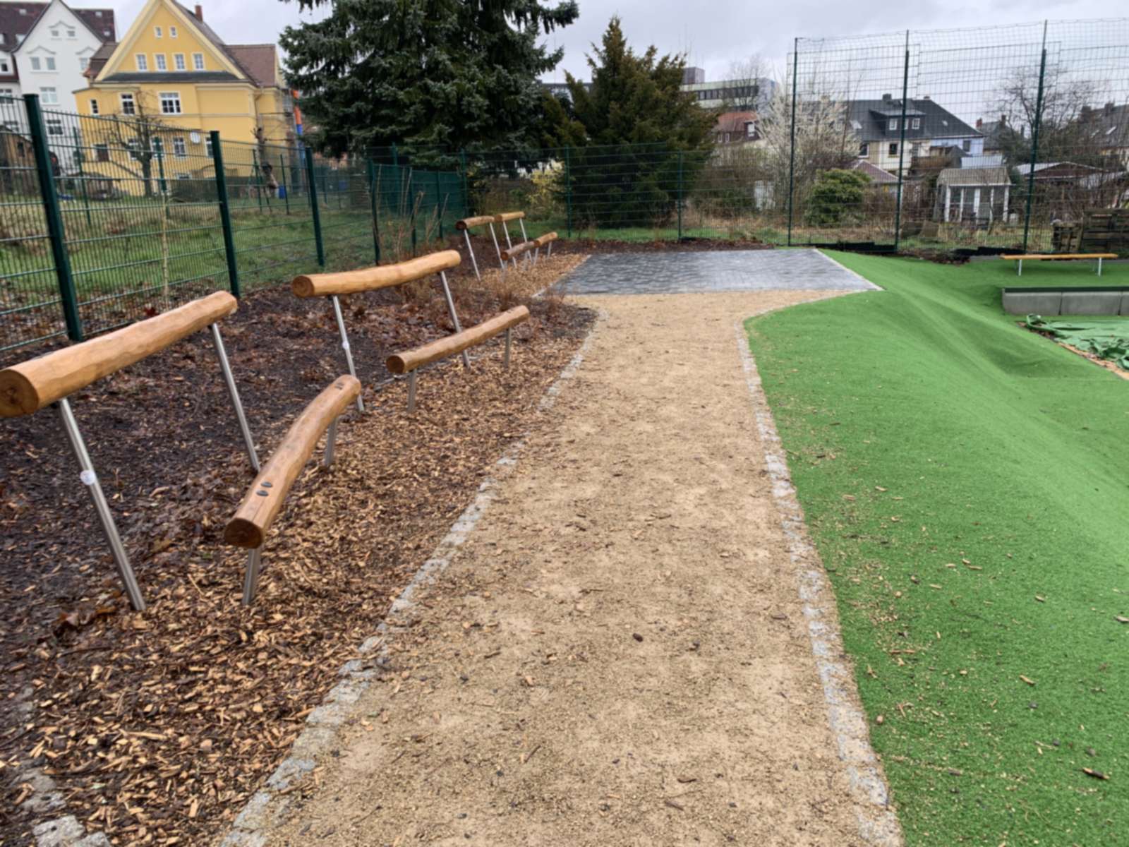 Wassergebundener Weg mit Balancierbalken aus Holz im Rindenmulchbeet am Rand des Sportfelds