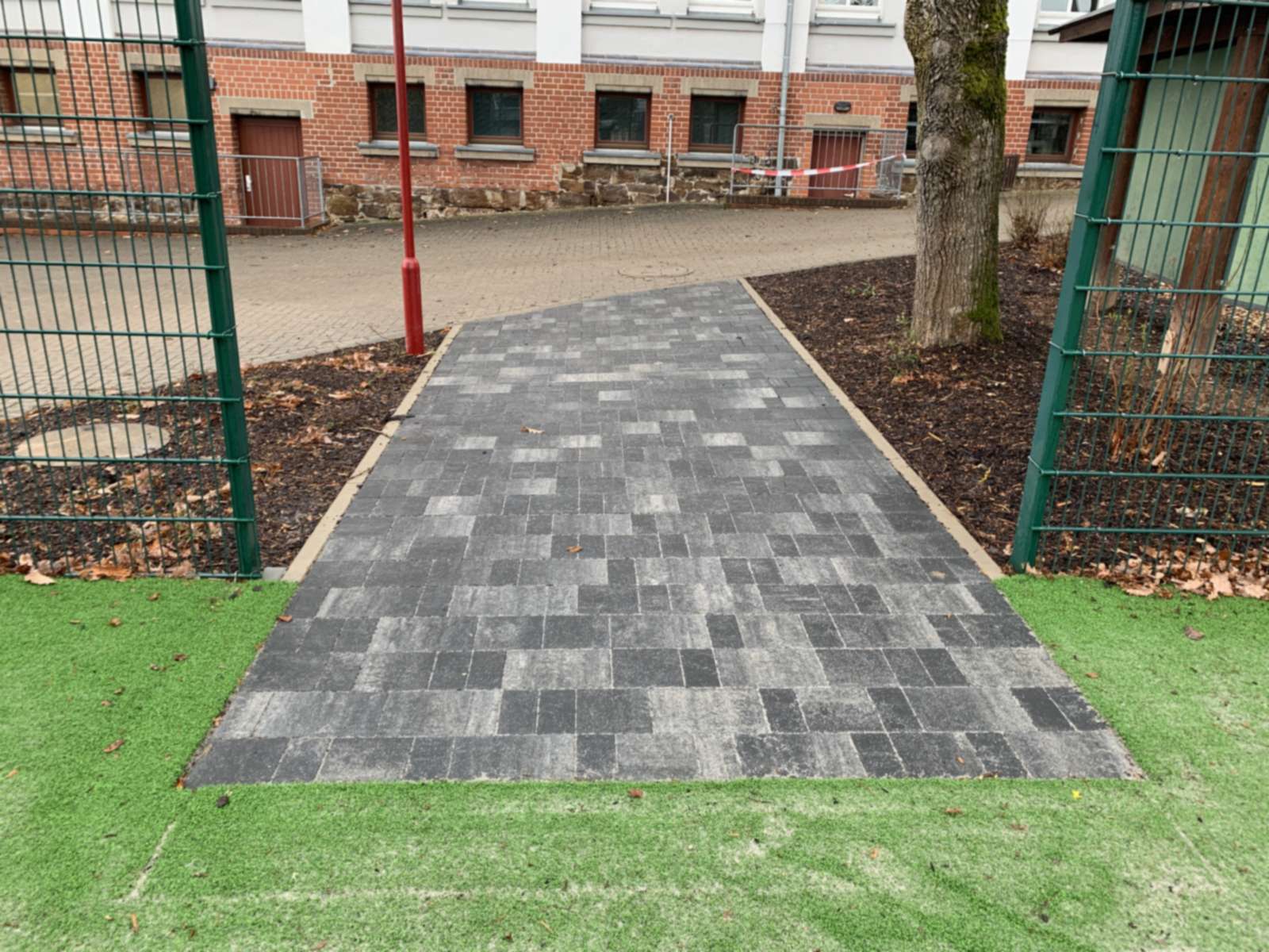 Gepflasterte Zuwegung vom Schulhof auf das Sportfeld zwischen Ballfangzaun und Baum