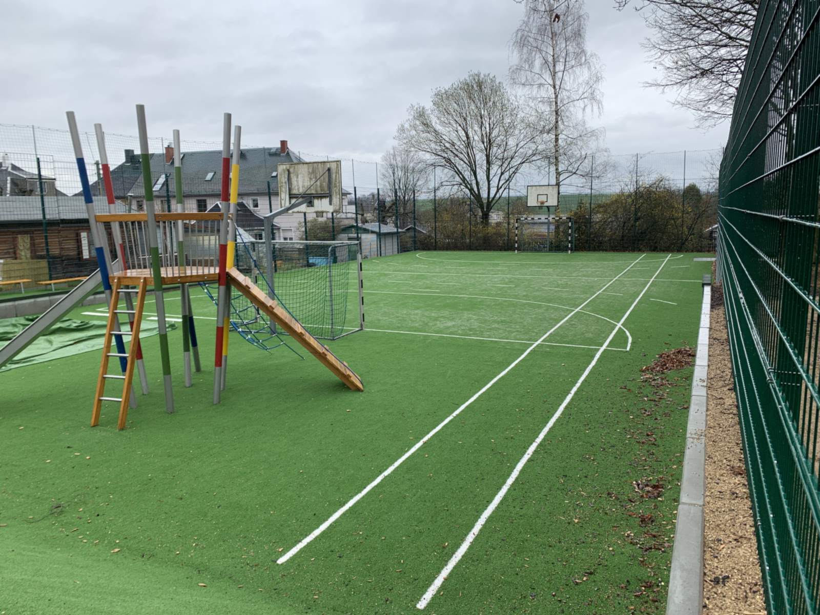 Sportfeld mit Kunstrasen und weißer Linierung, links eine Kletteranlage mit Rampe, dahinter Tore und Basketballkörbe