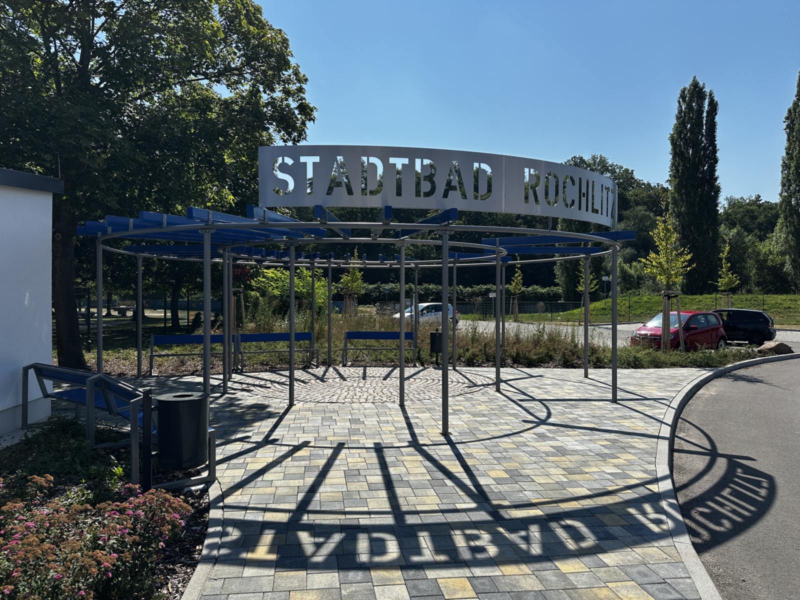 Überdachter Vorplatz mit Stahlpergola und dem Schriftzug Stadtbad Rochlitz, dessen Schatten auf das bunte Pflaster fällt