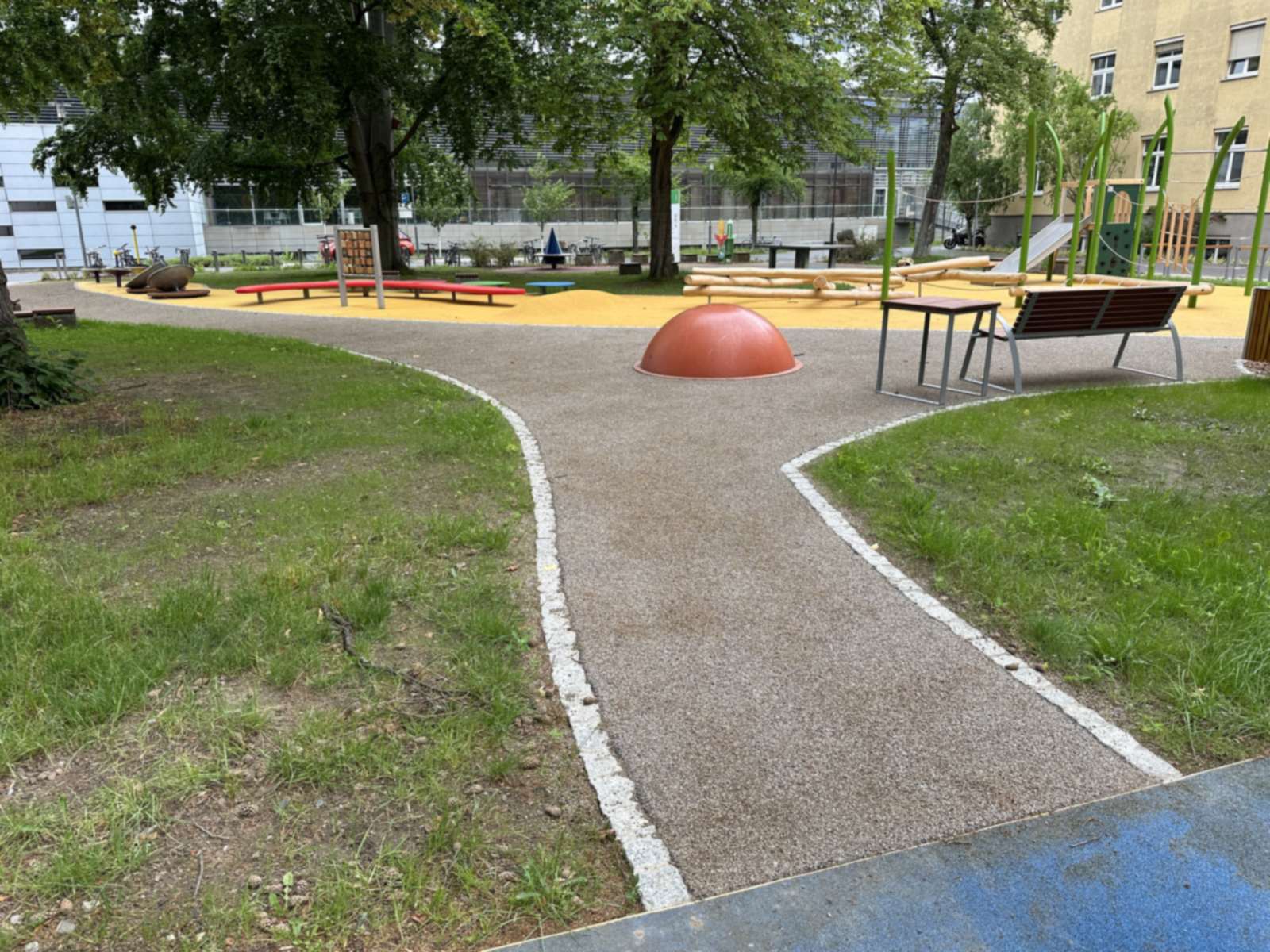 Geschwungener Weg mit wassergebundener Decke zwischen Rasenflächen, dahinter Spielhügel und gelber Fallschutzbelag