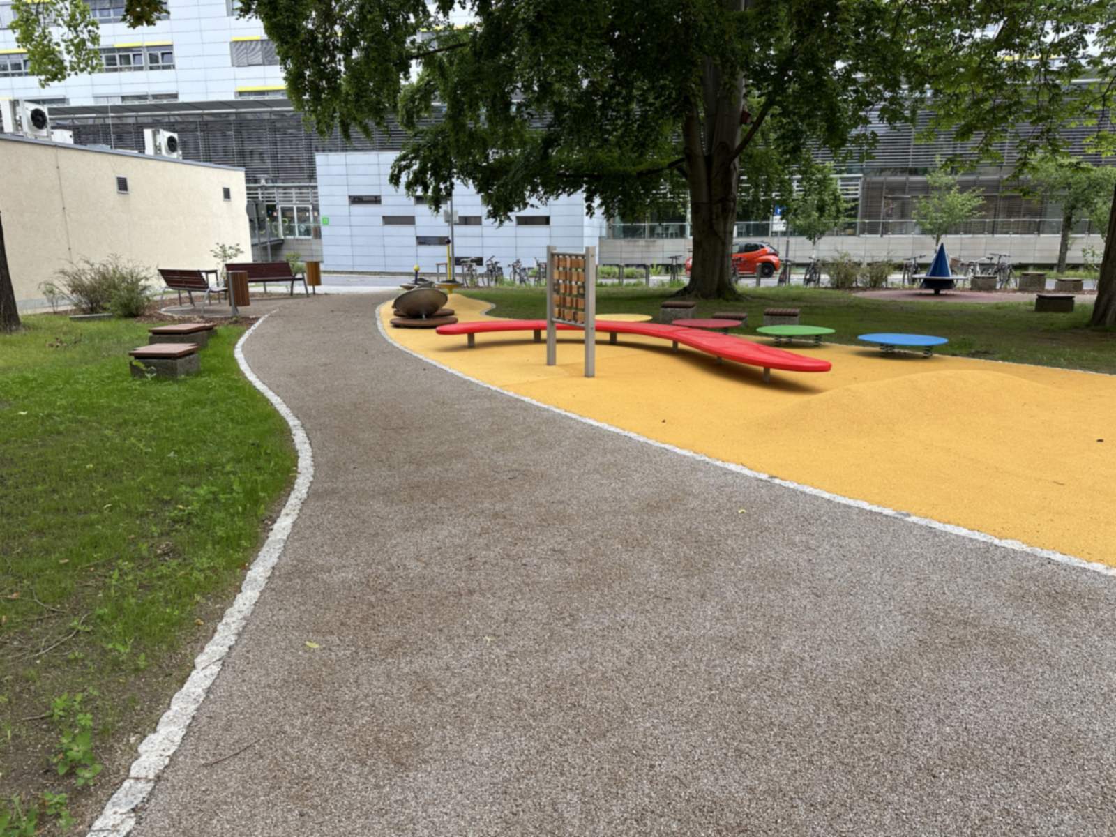 Weg entlang des gelben Spielbereichs mit roter Wellenbank und farbigen Drehtellern vor dem Klinikgebäude