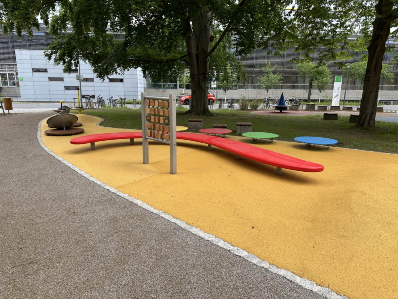 Spielbereich mit roter Wellenbank und Kletterwand auf gelbem Fallschutzbelag, daneben farbige Drehteller