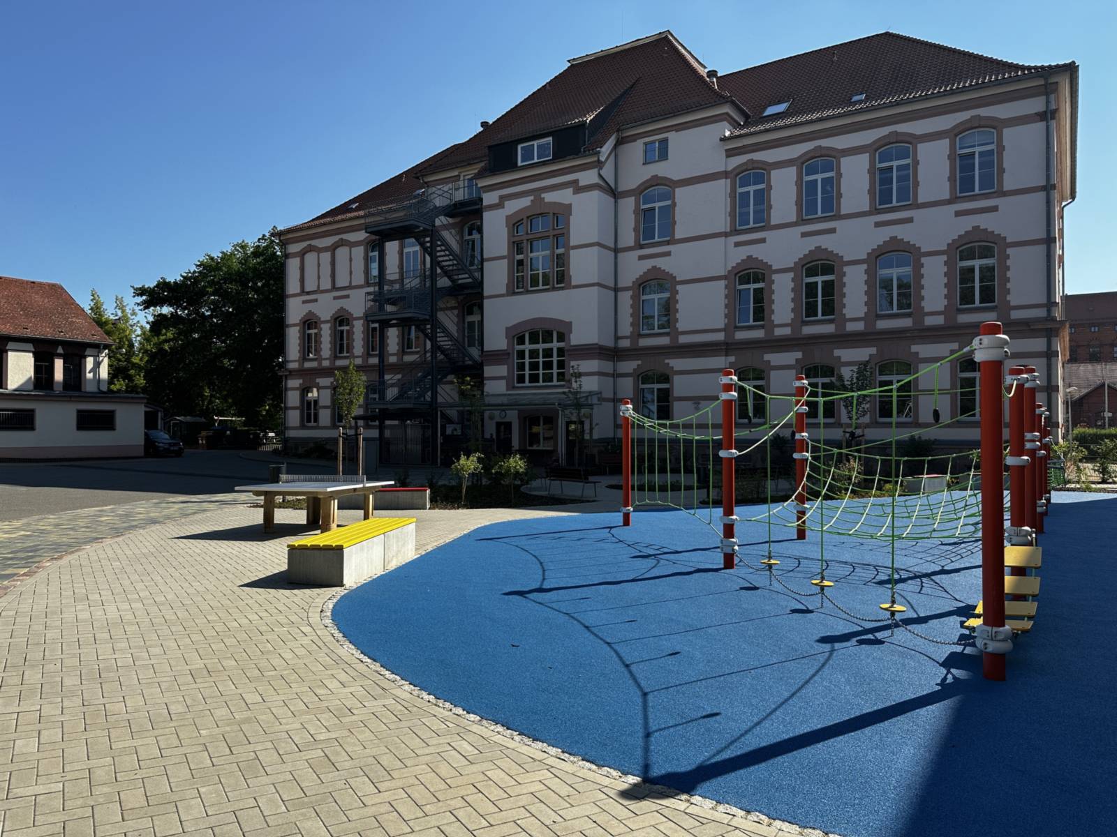 Schulhof mit blauem Fallschutzbelag und Hangelparcours vor dem historischen Schulgebäude, links eine Tischtennisplatte und eine gelbe Bank